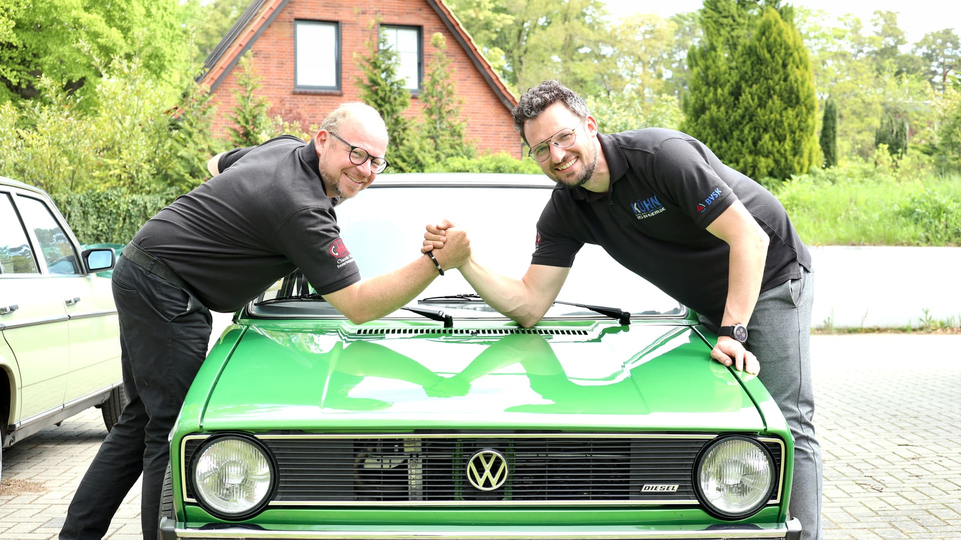 Karsten Kühn und Daniel Korfsmeier reichen sich über der Motorhaube eines grünen VW Golf die Hand